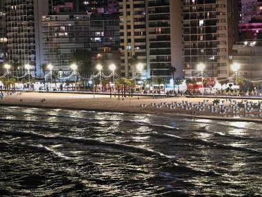 Sahilde yansımalar, sakin kıyı şeridi şehir parlaklığında ışıldayan su dokularıyla yıkanmış.