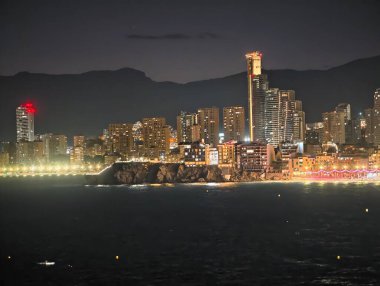 Yıldızlı gökyüzünün altında aydınlık bir sahil, parlak bir gece limanı manzarası, yükselen ışıklar ve sakin