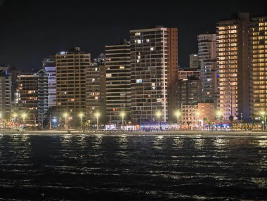 Neon ışıkları dalgalar üzerinde dans eder, şehir kıyı şeridi renkli neon ışıkları ve hareketli aktivitelerle aydınlatılır.