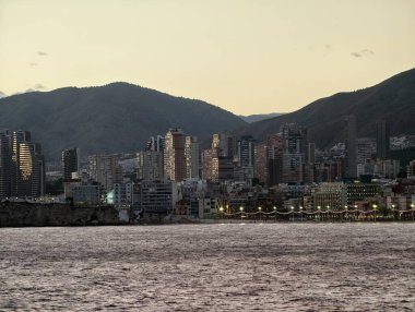 Akşam şehir manzarası, yuvarlanan tepelerin ve suyun üzerinde gün batımı, sakin bir alacakaranlık ve bastırılmış altın