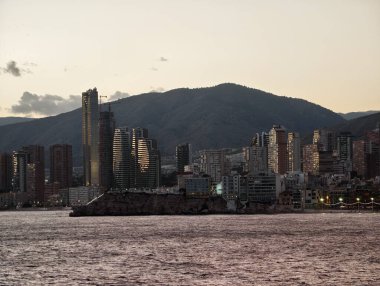 Dağın arka planında su yansımaları, feribot ve dağ ufkuyla huzurlu kıyı manzarası