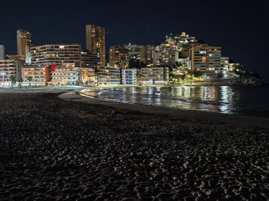Sahile bakan ufuk çizgisi, parlayan ışıklar, sakin gece manzaralı şehir ışıkları ıslak kumu yansıtıyor.