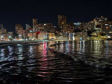 Suyun yanında parlak bir gece, parıldayan gökdelenler ve ışıldayan suyla dolu canlı bir akşam atmosferi.