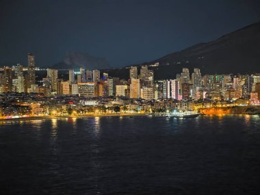 Şehrin ufuk çizgisi parlıyor, geniş deniz manzarası parlayan kuleler ve sisli gece yansımaları içeriyor.