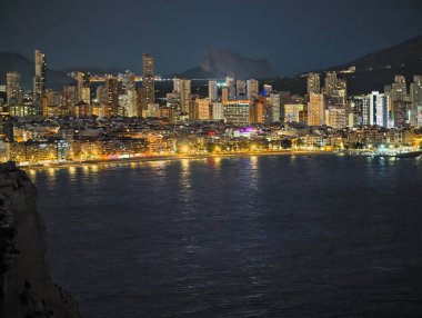 Alacakaranlıktaki hareketli limanda parlak binalar ve manzaralı dağ manzarası