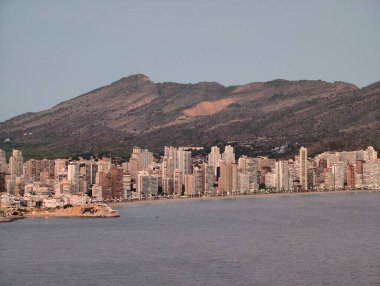 Şehir manzarasının arkasındaki engebeli dağ sıraları, kümelenmiş kuleleri ve uzak dağları olan güneşli kıyı şeridi.