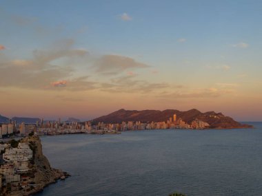 Gün batımında huzurlu deniz manzarası, uzak dağ siluetleri ve yumuşak gökyüzü yansıması olan sessiz körfez.