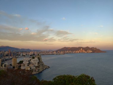 Kıyı günbatımı manzarası, alacakaranlık siluetleri sakin okyanusun üzerindeki aydınlık bulutlarla nazikçe örtülmüş.