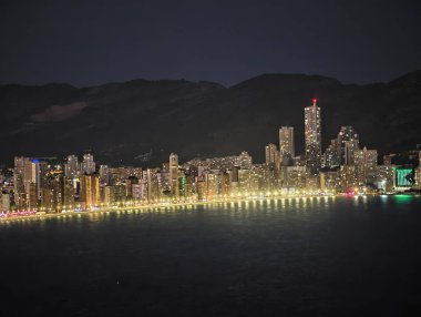 Gece vakti, ışıklandırılmış rıhtım manzarası ve yüksek gökdelenleri ile dağ zeminine karşı