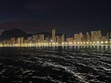 Canlı deniz kenti, hareketli dalgaların üzerindeki parlak şehir ışıkları canlı gece deniz manzarası yaratır.