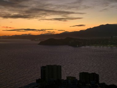 Akşam liman manzarası, siluetli tepeler ve yükselen fenerlerle sakin bir akşam.