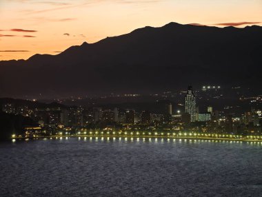 Alacakaranlıktaki huzurlu sahil şehrinde rıhtım ve dağ hatları boyunca parlak ışıklar var.