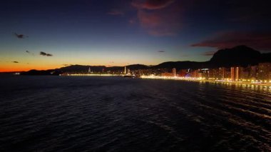 Sahil manzarası, alacakaranlık ufku parıldayan dalgalar ve aydınlatılmış sahil manzarası.