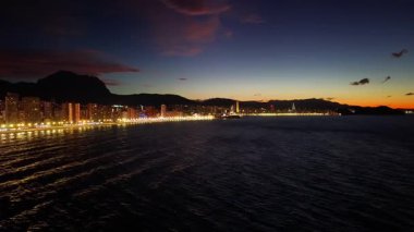 Alacakaranlık, sakin kıyı şeridini parlak ışıklarla, yumuşak dalgalarla ve alacakaranlıkta parlak ufuk çizgisiyle boyar.