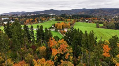 Hava manzaralı ağaçlar ön planda, canlı turuncu gölgelikler uzak tarım arazilerini ve dolambaçlı yolları çerçeveliyor.