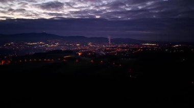 Bulutlu gökyüzünün altında aydınlık mahalleler, parlayan şehir sokakları ve endüstriyel manzaralar.