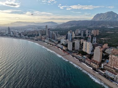 İHA 'dan gelen gündoğumu sahili manzarası geniş kıyı şeridi, kümelenmiş kuleler ve durgun turkuaz deniz.