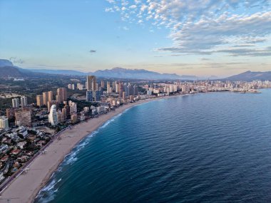 Yükselen ufuk çizgisi ve berrak gökyüzü altında geniş kum şeridi olan eğimli körfez sahili, görüntü deniz kenarını vurguluyor