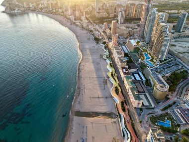 Kıvrımlı körfezin üzerinde hava gündoğumu ve boş plaj, turkuaz su altın ışığı yansıtıyor, modern kuleler hizasında.