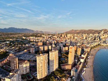 Hava sahili ufku alacakaranlıkta uzun kumlu kıyı, tatil köyü kuleleri, hareketli gezinti güvertesi, sıcak gökyüzü yansımaları