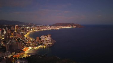 Aydınlatılmış Körfez üzerindeki canlı gece hayatı, Kuleler ve hareketli caddelerle kaplı hilal plajının havadan görünüşü,