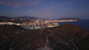 Parlayan kuleleri ve rıhtım ışıkları, kıvrımlı yamaçları olan Aydınlatılmış Sahil Şehri Gece Havası Görünümü