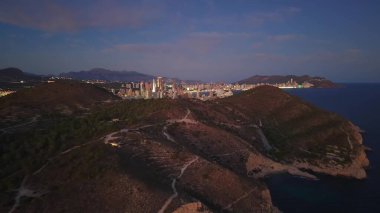 Uçan Alacakaranlık Kıyı Tepeleri 'nin üzerinde. Körfez boyunca parlayan şehir ışıkları ve kıvrımlı uçurumlar.