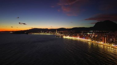 Parlayan Deniz Yansımaları ve Dağ Arkaplanı ile Alacakaranlıkta Kumsalda Hava Aydınlatılmış Gezinti Alanı,