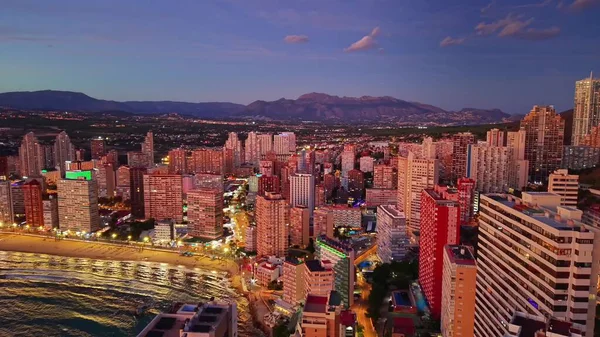 Alacakaranlık Şehri Kıyı şeridi boyunca, Aydınlatılmış Gezinti Alanı ve Meşgul Sahil Binaları Mor Gökyüzü Altında Parlıyor,