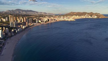 Sakin su ve berrak gökyüzü, plajın uzun kıvrımı, şehir blokları ve uzak mesafeli