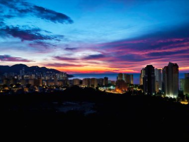 Skyline üzerinde canlı mavi saat, kıyı kuleleri ve neon ışıkları, dramatik katmanlı bulutlar, yansıtıcı deniz.