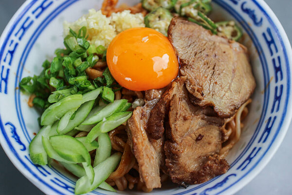 Thai-style Sichuan Red Pork Noodle bowl featuring an uncooked egg yolk, char siu pork slices, and cucumber. This rich and savory Thai street food offers a delicious blend of Asian flavors and textures.