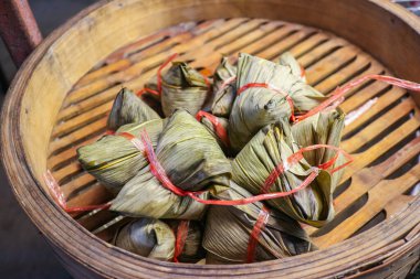 Pile of Sticky Rice Dumplings (Bajang or Zongzi) wrapped in banana leaves and tied, displayed in an old bamboo steamer. Classic Thai/Chinese street food.