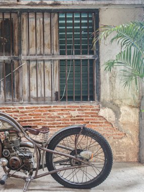 Eski tuğla duvar arka planında klasik motosiklet. Grunge desenli, eski pencere ızgaralı ve tropikal palmiye yapraklı kırsal bir sokak manzarası. Seyahat, tarih veya kentsel çürüme kavramları için ideal.