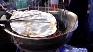 Parlayan kırmızı kömür üzerinde genişleyen dev bir Tayland pirinç krakerinin yoğun yakın çekimi. Becerikli bir sokak satıcısı, atıştırmalıkları titizlikle çeviriyor. Otantik Asya gastronomisinin ısısını ve çıtır çıtır dokusunu yakalar..