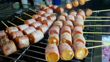 Sıcak kömür közlerinin üzerine peynirli sosis sarılmış domuz pastırması şişleri. Yerel bir marketteki geleneksel Tayland sokak yiyeceklerinin canlı atmosferini ve iştah açıcı görüntülerini yakalıyorum..