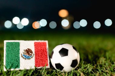 Meksika ulusal bayrağı, geleneksel bir futbol topunun yanına yerleştirilmiş aydınlık yeşil stadyum çimlerinin üzerine gece. Dramatik bokeh ışıkları ulusal gurur, birlik ve uluslararası spor müsabakalarını temsil eden küresel futbol turnuvası atmosferini geliştirdi.