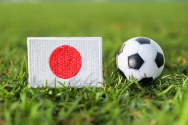Japonya ulusal bayrağı, Asya futbol kültürünü ve uluslararası yarışma konseptini temsil eden bulanık saha arkaplanlı yumuşak güneş ışığıyla aydınlatılmış canlı futbol stadyumu çimlerinin üzerinde klasik bir futbol topunun yanında..