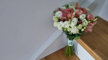 Beyaz ve pembe çiçeklerin yer aldığı yuvarlak bir düğün buketi ahşap basamaklarda duruyor. Bukette beyaz güller ve yeşil yapraklı pembe Alstromerya yer alıyor..