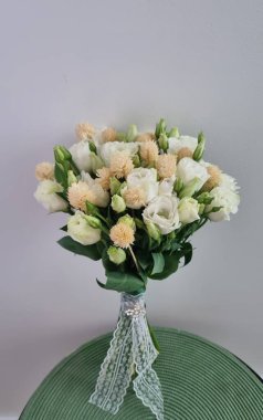 Başta Eustoma (Eustoma grandiflorum) olmak üzere beyaz ve krema çiçeklerinin yer aldığı düğün buketi, soluk şeftali çiçekleriyle aksanlıdır. Dantele sarılmış, sapları dekoratif bir yayla sabitlenmiş..
