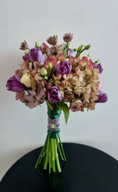 Menekşe renginde yuvarlak bir düğün buketi, ortanca ve lalelerden yapılmış, siyah bir masanın üzerinde beyaz bir arka planda duruyor. Gelin tasarımı ve dekorasyonu için zarif bir çiçek aranjmanı.