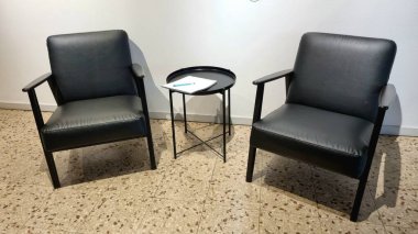 Masaların arasında iki siyah koltuk, içinde bir defter ve tavan arasında bir kalem olan siyah demir bir masa.