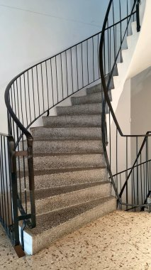 Girişin iç tarafındaki gri basamaklı metal spiral merdiven. Tırabzanlar yukarı doğru kıvrımlı, siyah metalden yapılmış. Duvarlar yumuşak gri bir tonla boyanmış, modern bir atmosfer yaratıyor.