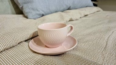 Uyumlu bir fincan pembe çay fincanı çizgili yatağın üzerinde duruyor. Yatak örtüsü, arka planda açık mavi bir yastıkla tamamlanmış, nötr tonlarda ince çizgilerden oluşan bir desen içeriyor..