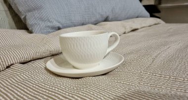 Çizgili Yatakta Sıcak Sabah Çay Fincanı. Yatağında aromatik kahve olan beyaz fincan, konfor konsepti ve ev rahatlığı atmosferi.