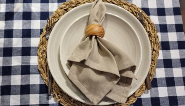 Zarif ev sofra takımı, mavi ve beyaz çekli klasik masa örtüsü, hasırdan yuvarlak masa ve bej tabaklar. Bir peçete bir tabak dokusunu ahşap bir yüzük ve cupronickel aletlerle süslüyor.