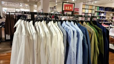 Beyaz, mavi, bej ve yeşil renkli erkek gömlekleri bir butikte siyah metal satış rafında düzgünce düzenlenmiştir. Perakende ortamda parlayan ahşap bir zemin ve tepeden aydınlatma yer alıyor..