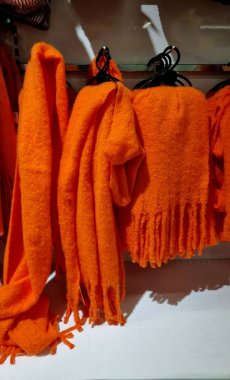 Püsküllü turuncu eşarplar bir mağazadaki askılarda sergileniyor, metal bir rafta düzgünce düzenleniyor. Eşarplar kabarık ve dokulu.,