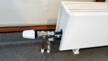 Termostatik valf takılı, ayarlanabilir ayarları olan beyaz radyatör. Vana metal boruya bağlı, kısmen tahta bir etek tahtasıyla kaplı. Radyatör benekli gri bir zeminde duruyor.