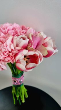 Yumuşak pembe karanfil buketi ve karanlık dairesel bir yüzeyde laleler. Karanfiller yumuşak pembe renklere sahip fırfırlı taç yaprakları sergilerken, laleler pembe ve beyaz renklere sahip pürüzsüz yapraklara sahipler..